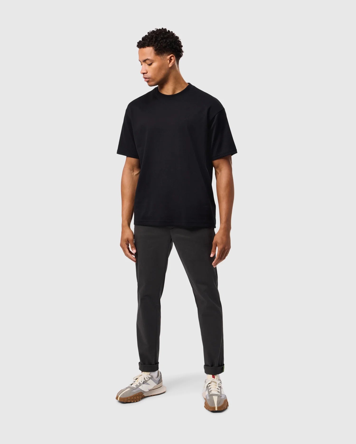 A man wearing a Psycho Bunny MENS DUKE OVERSIZED TEE - B6U742D200 in heavyweight Pima cotton, dark pants, and white sneakers stands against a plain light gray background. He looks down to his left with hands relaxed at his sides. His outfit is casual and minimalistic.