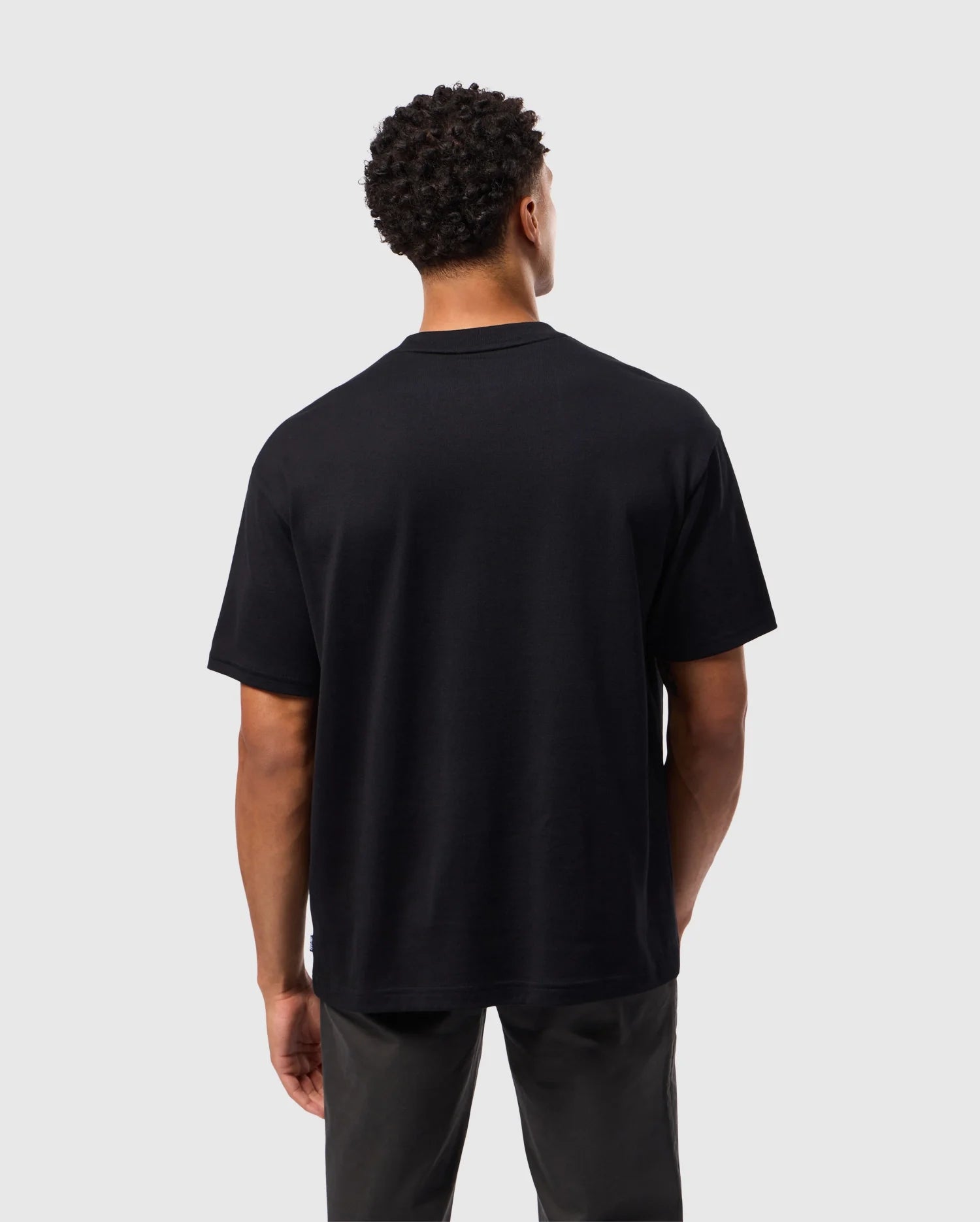 A person with short curly hair is standing and facing away from the camera, showcasing the relaxed fit of the Psycho Bunny MENS DUKE OVERSIZED TEE (B6U742D200) in plain black heavyweight cotton, paired with dark gray pants. The background is plain and light-colored.