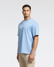A man with short curly hair stands against a plain background, wearing a light blue Psycho Bunny MENS DUKE OVERSIZED TEE - B6U742D200 and beige pants. The relaxed fit complements his neutral expression as he looks directly at the camera.