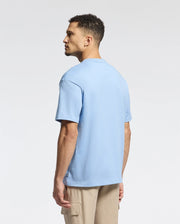 A man with short curly hair stands slightly turned wearing a Psycho Bunny MENS DUKE OVERSIZED TEE in light blue Pima cotton and beige cargo pants, effortlessly complementing the plain white background.