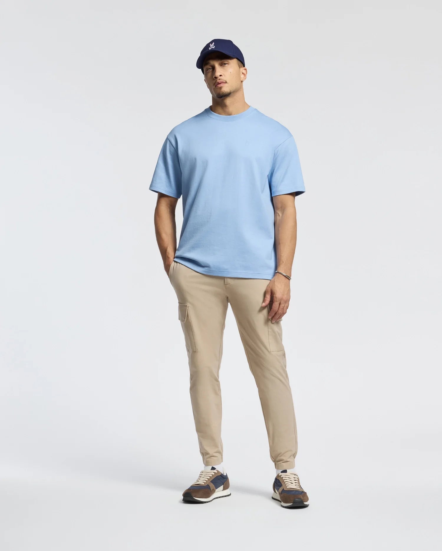 A man in a Psycho Bunny Men's Duke Oversized Tee (B6U742D200)—a blue heavyweight jersey—teams it with beige pants, a dark baseball cap, and sneakers, casually posing against a plain white background.