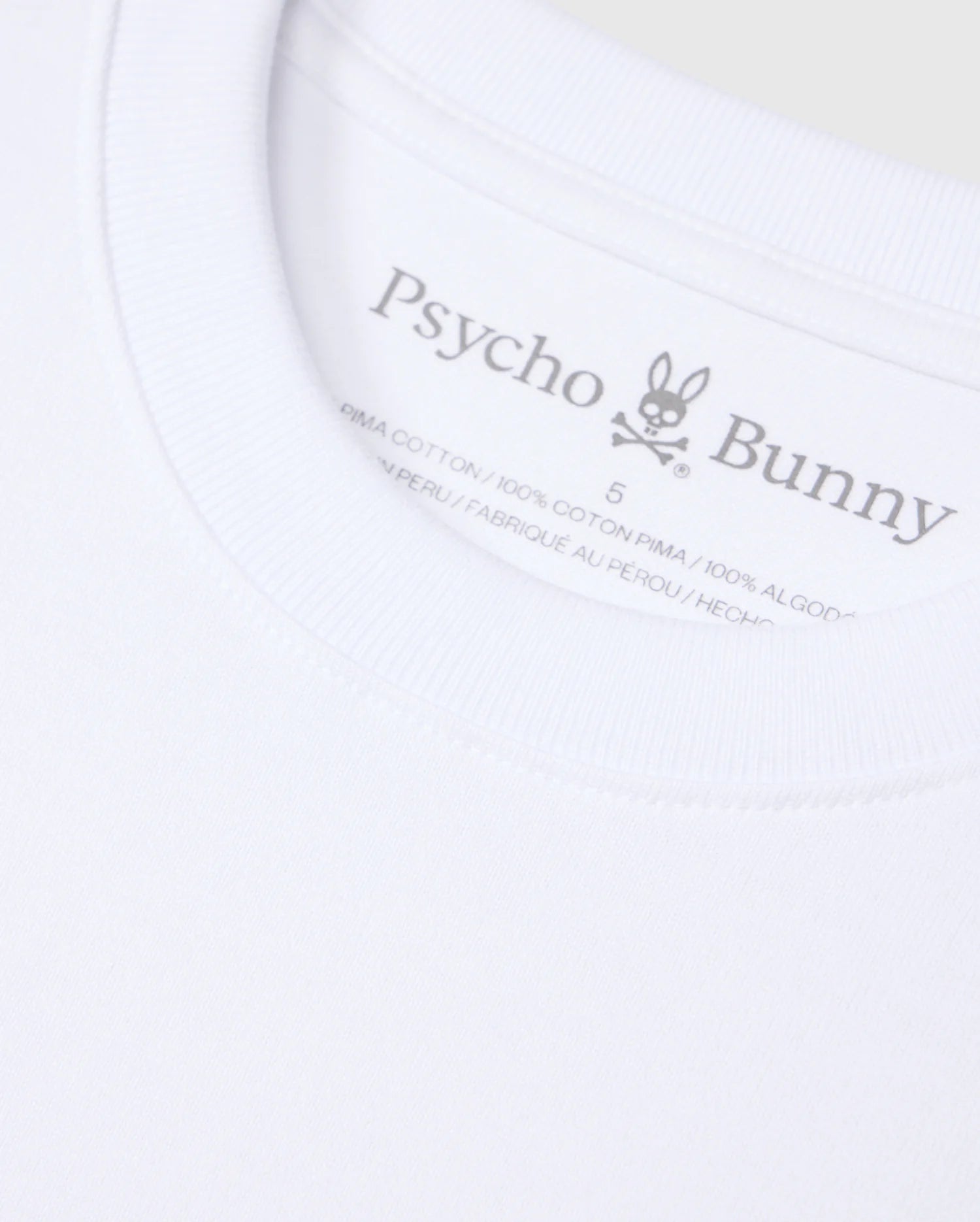Close-up of a white Psycho Bunny t-shirt label. The label displays the brand name "Psycho Bunny," a rabbit logo with crossbones, and indicates the shirt is made from 100% heavyweight Pima cotton and manufactured in Peru. The background of the image is the solid pattern fabric of this relaxed fit MENS DUKE OVERSIZED TEE - B6U742D200.