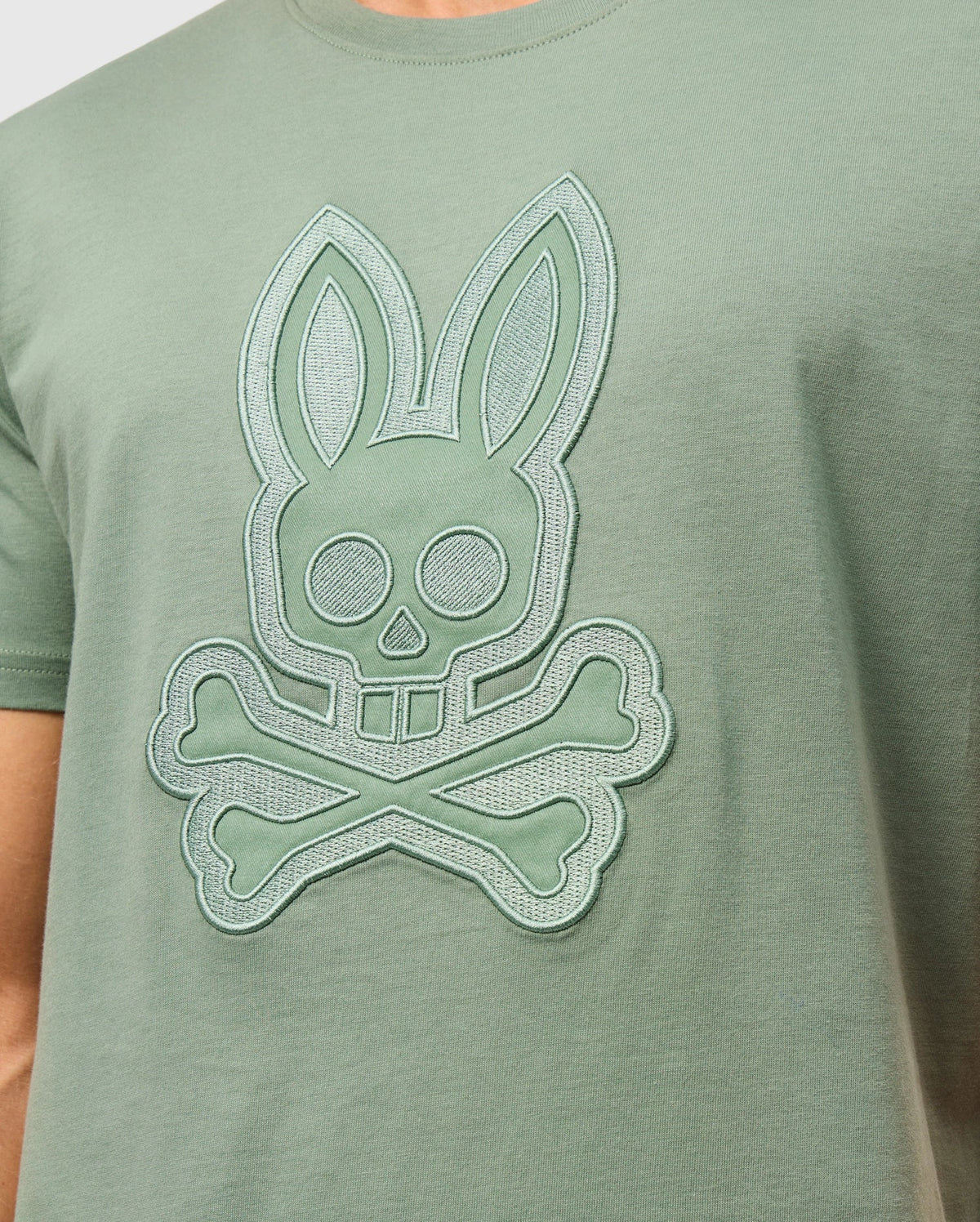 A person wearing the Psycho Bunny MENS MALTON GRAPHIC TEE - B6U795D200, a green Peruvian-crafted t-shirt featuring an embroidered rabbit skull with crossbones design. The graphic includes large circular eyes and two long ears, creating a playful yet slightly edgy look. This comfortable tee is made from soft Pima cotton.