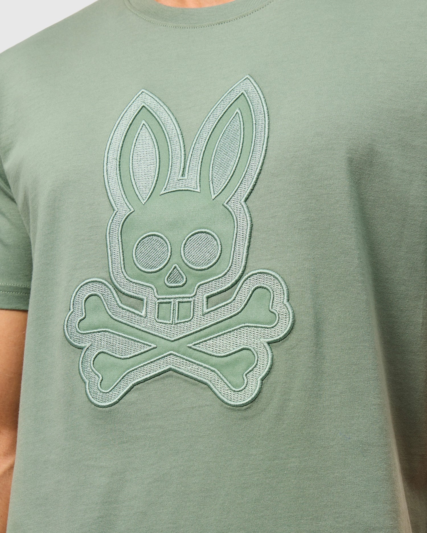 A person wearing the Psycho Bunny MENS MALTON GRAPHIC TEE - B6U795D200, a green Peruvian-crafted t-shirt featuring an embroidered rabbit skull with crossbones design. The graphic includes large circular eyes and two long ears, creating a playful yet slightly edgy look. This comfortable tee is made from soft Pima cotton.