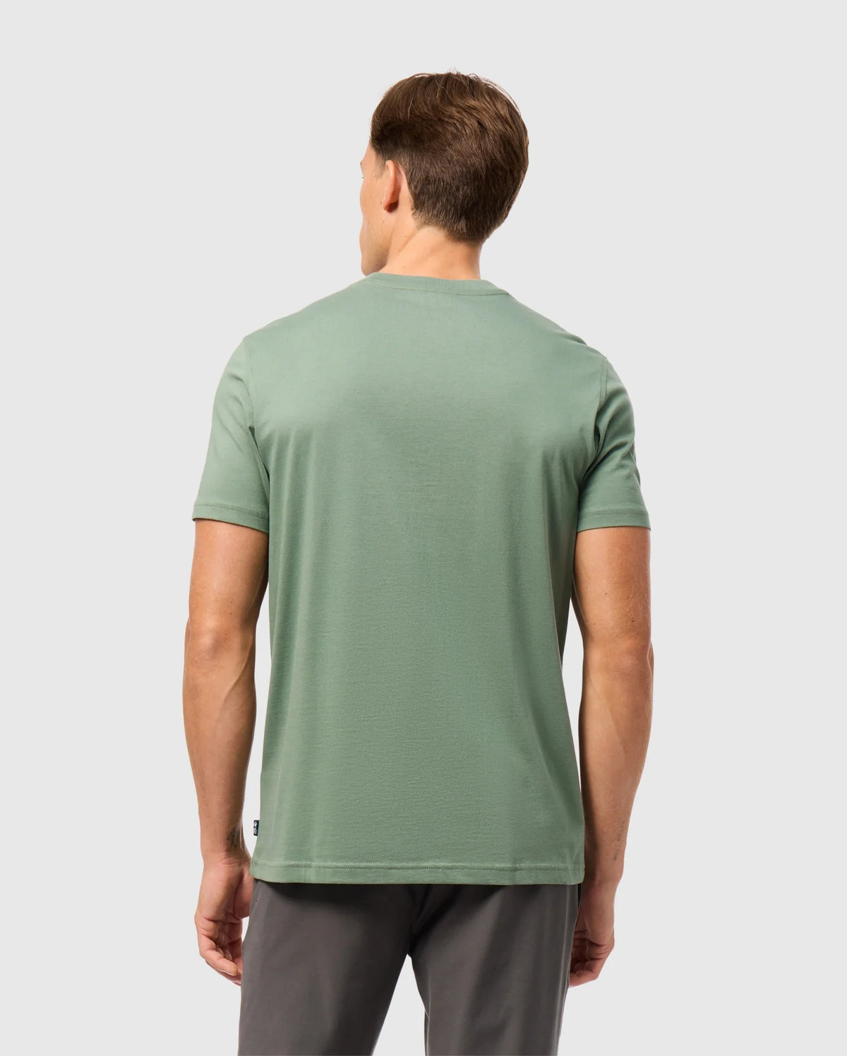 A man with short brown hair stands with his back to the camera, wearing a light green Psycho Bunny MENS MALTON GRAPHIC TEE - B6U795D200 and dark gray pants against a plain light gray background.