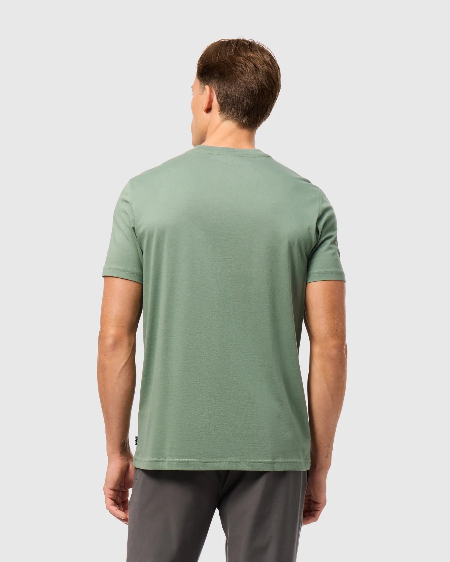 A man with short brown hair stands with his back to the camera, wearing a light green Psycho Bunny MENS MALTON GRAPHIC TEE - B6U795D200 and dark gray pants against a plain light gray background.
