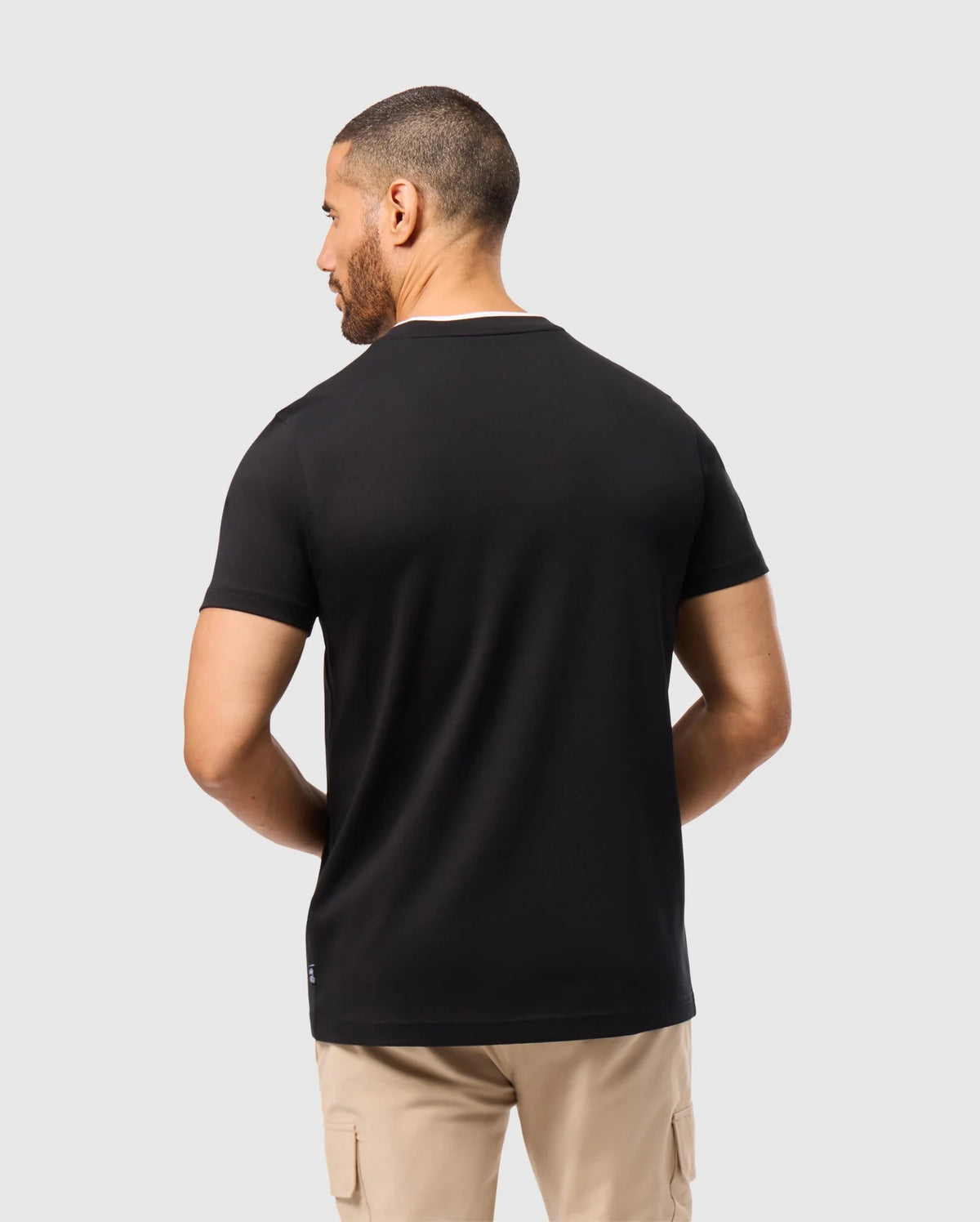 A person with short hair wears the Psycho Bunny MENS CAMPO POCKET TEE - B6U801D200 in black, crafted from pima cotton, paired with beige pants, and stands facing away from the camera against a plain gray background.
