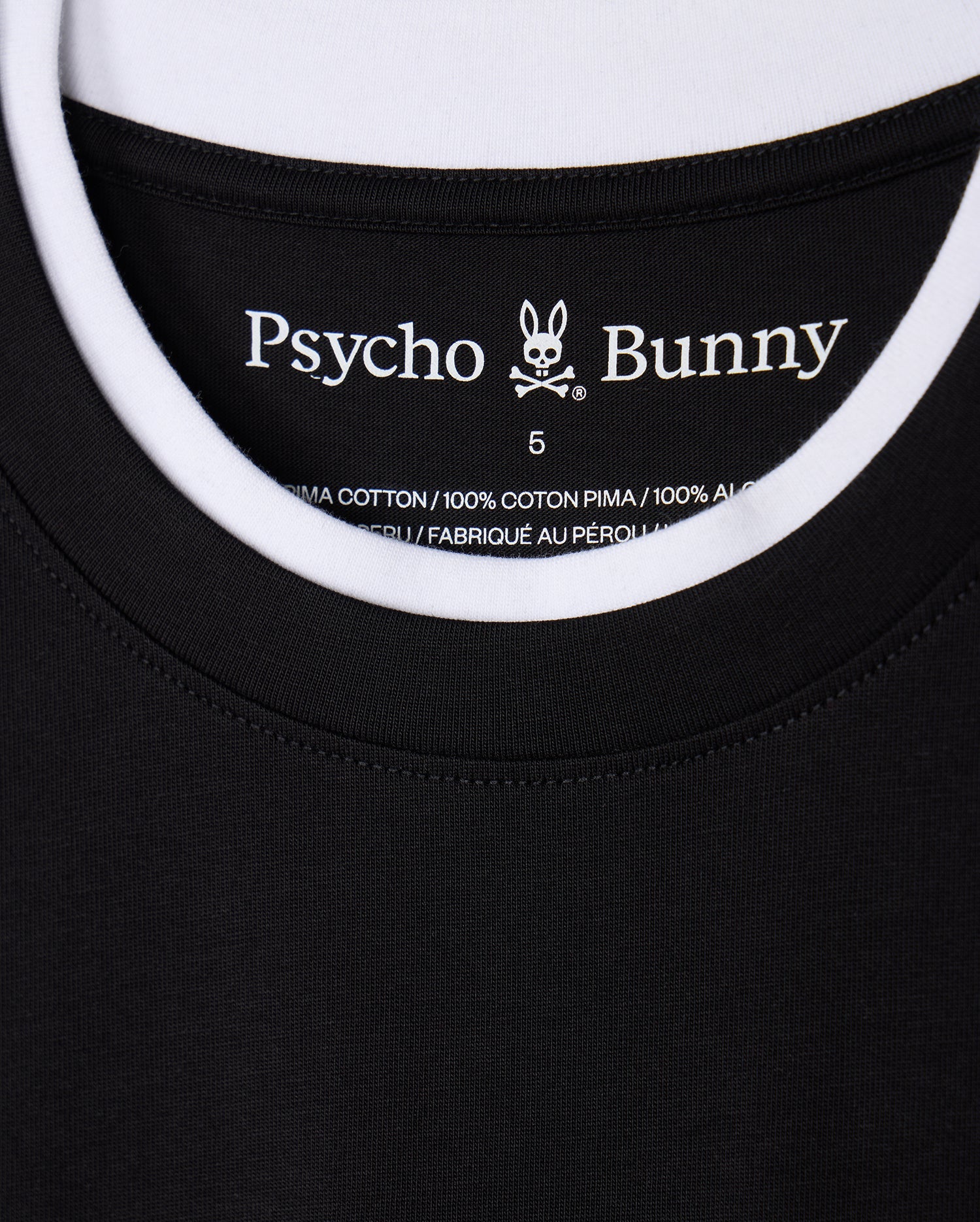 Close-up of a black and white double collar T-shirt, identified as the "MENS CAMPO POCKET TEE - B6U801D200," featuring a label with "Psycho Bunny" text and a bunny logo. The label states that it is made of 100% Pima cotton material and manufactured in Peru.