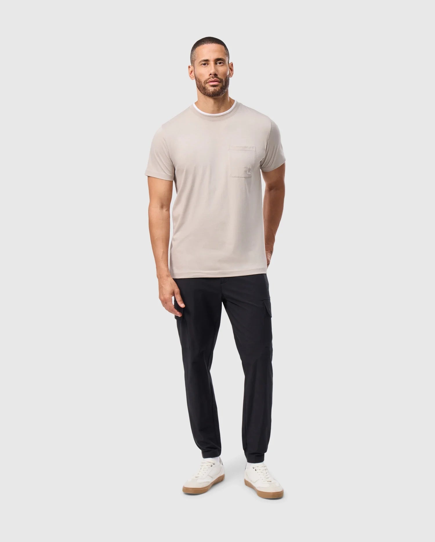 He stands against a plain background wearing the Psycho Bunny MENS CAMPO POCKET TEE - B6U801D200 in beige, paired with black pants. He has short hair, a beard, and is sporting white sneakers. His hands hang relaxed by his sides.