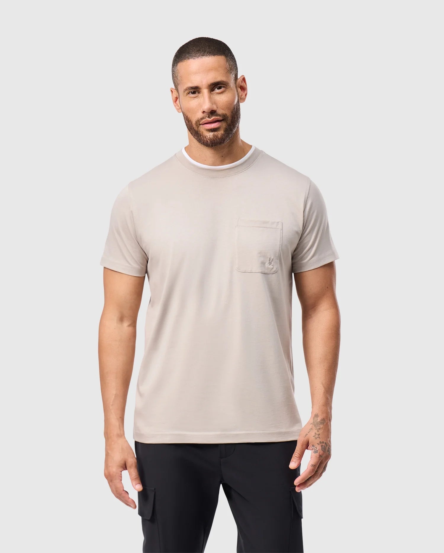 A man with short hair and a beard wears a Psycho Bunny MENS CAMPO POCKET TEE in light gray, made from soft pima cotton. The tee pairs effortlessly with his black pants as he stands against a plain white background.