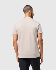A man wearing the Psycho Bunny MENS CAMPO POCKET TEE in light beige, made from soft pima cotton, and black pants stands with his back to the camera against a plain gray background.