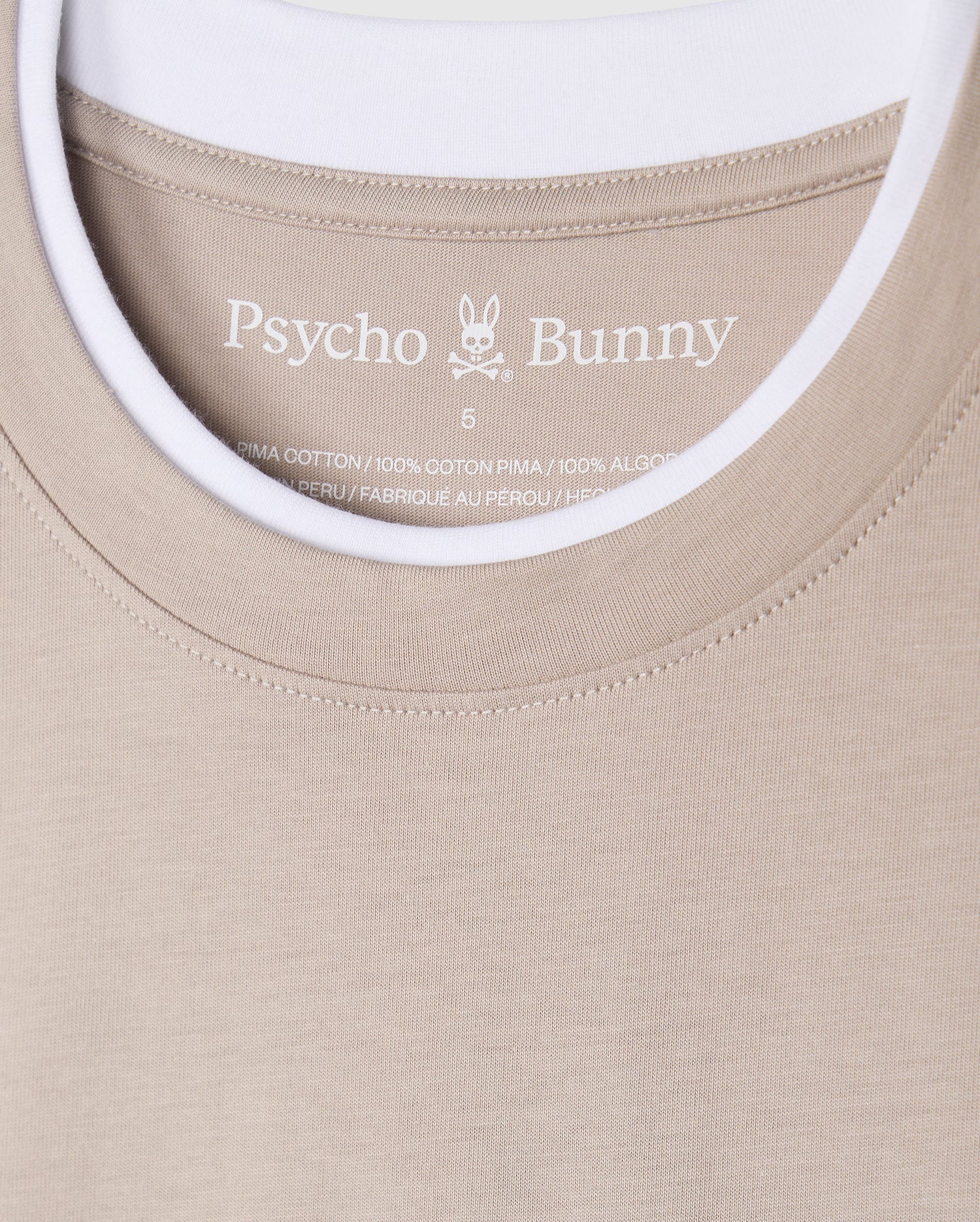 The Psycho Bunny MENS CAMPO POCKET TEE (B6U801D200) is a beige pocket tee showcasing the bunny logo, crafted from 100% Pima cotton in Peru. It includes printed label details inside and features a stylish double collar revealing a white t-shirt collar underneath.