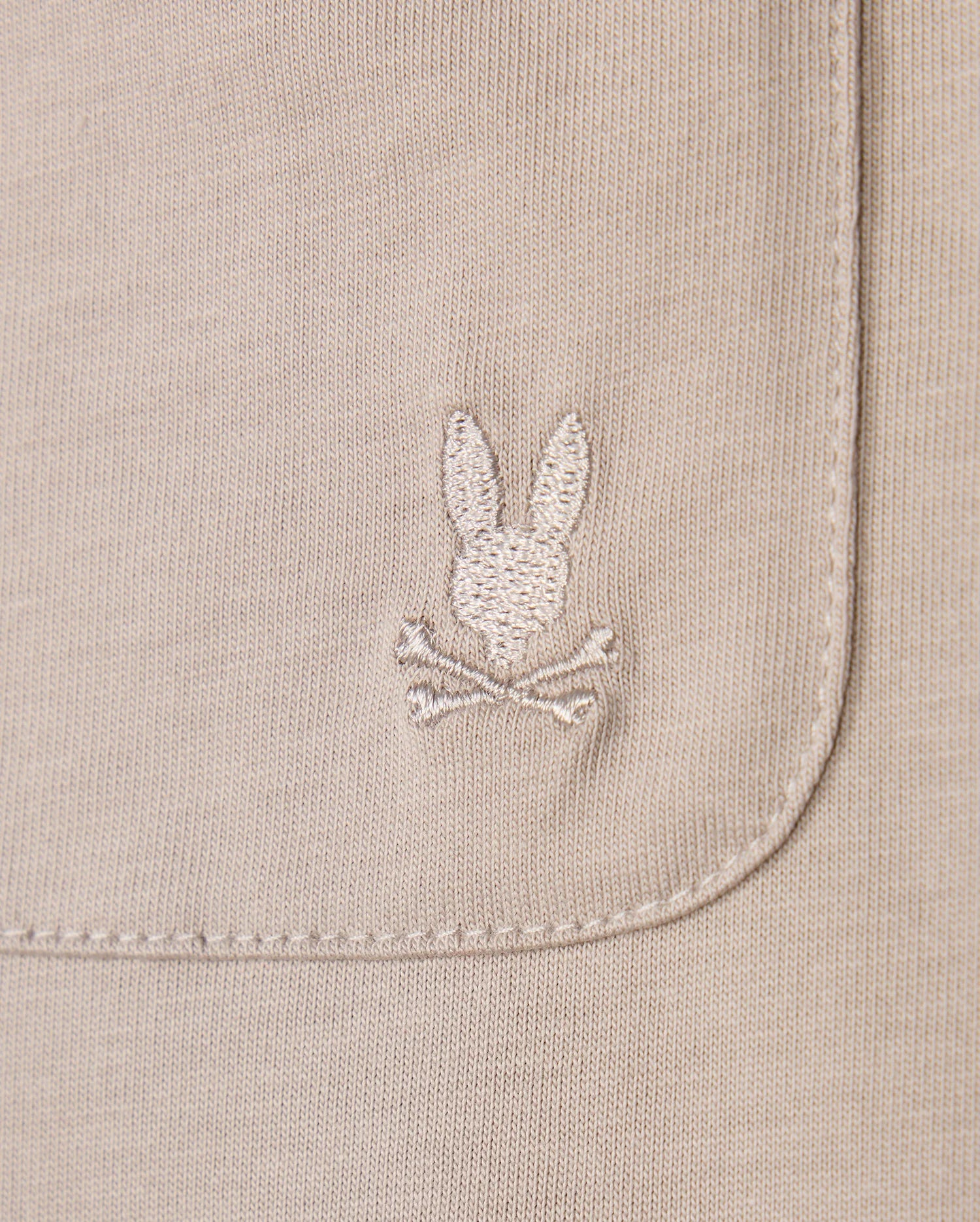 Close-up of the Psycho Bunny MENS CAMPO POCKET TEE - B6U801D200 in beige pima cotton, showcasing an embroidered rabbit's head and crossbones on the pocket. The texture and detailed stitching are clearly visible.