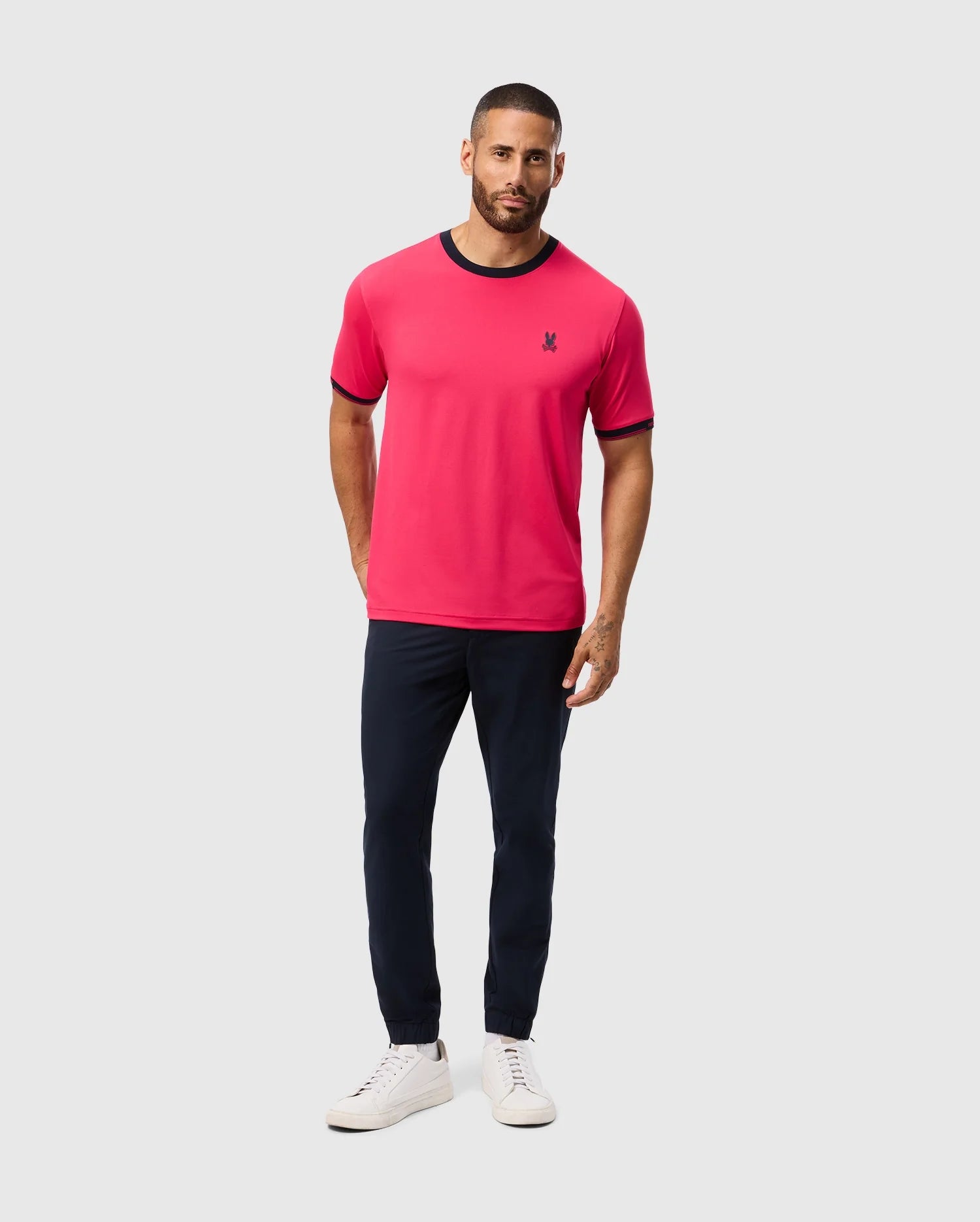 A man sports a Psycho Bunny MENS EDMONDS SPORT TEE (B6U857D200) in bright pink, featuring a small chest logo. He pairs it with navy pants and white sneakers. The tee's quick-drying, antimicrobial fabric suits the light gray backdrop as he stands with hands in pockets and a neutral expression.