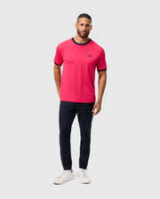 A man sports a Psycho Bunny MENS EDMONDS SPORT TEE (B6U857D200) in bright pink, featuring a small chest logo. He pairs it with navy pants and white sneakers. The tee's quick-drying, antimicrobial fabric suits the light gray backdrop as he stands with hands in pockets and a neutral expression.
