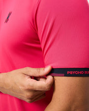 A person wears the MENS EDMONDS SPORT TEE by Psycho Bunny, featuring a small logo on the chest. The pink short-sleeve tee is made from antimicrobial fabric. They hold a sleeve edge with a black band reading "PSYCHO BUN" in red text.