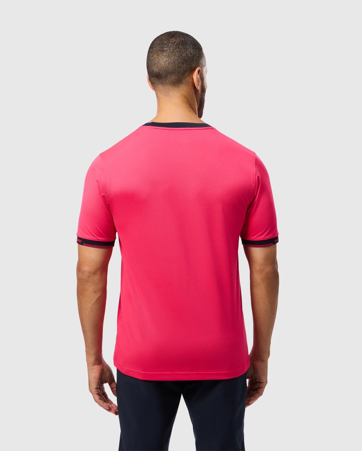 A person with short hair faces away from the camera, showcasing the Psycho Bunny MENS EDMONDS SPORT TEE - B6U857D200, a bright red tee with dark trim on sleeves and collar. Made of quick-drying, antimicrobial fabric, it pairs well with their dark pants against a plain light gray backdrop.
