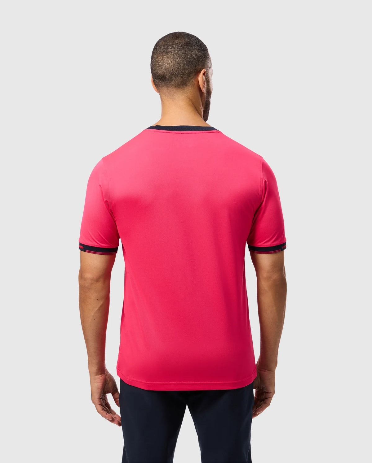 A person with short hair faces away from the camera, showcasing the Psycho Bunny MENS EDMONDS SPORT TEE - B6U857D200, a bright red tee with dark trim on sleeves and collar. Made of quick-drying, antimicrobial fabric, it pairs well with their dark pants against a plain light gray backdrop.