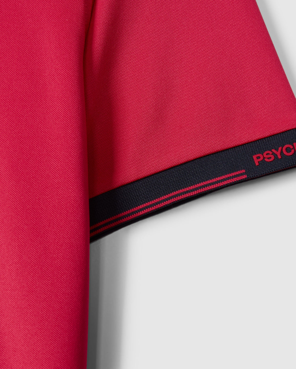 Close-up of a red Psycho Bunny MENS EDMONDS SPORT TEE sleeve, featuring a black edge with pink stripes and partially visible "PSYC" text. The textured, antimicrobial fabric enhances functionality against the light gray background.