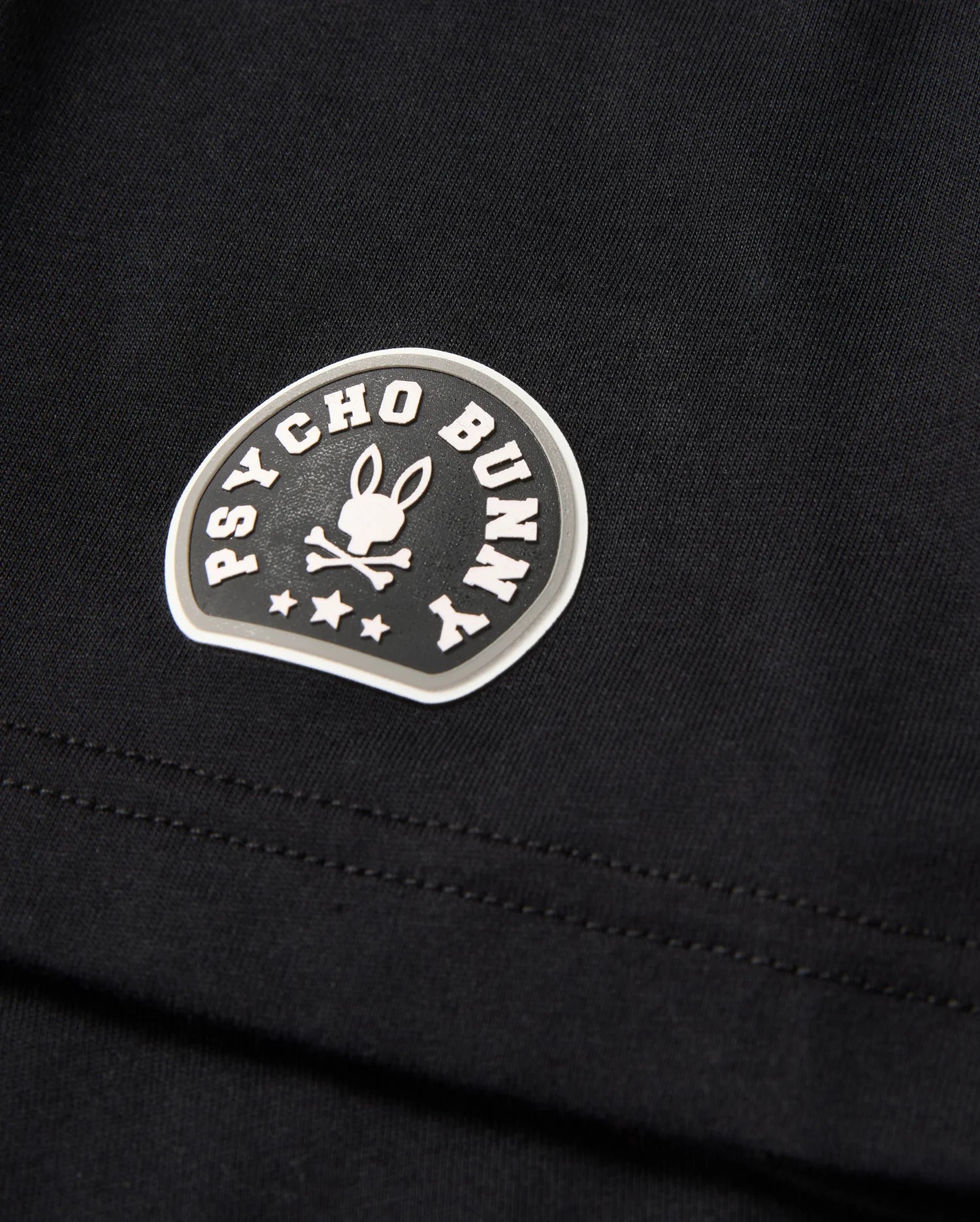 Close-up of the MENS ZACK GRAPHIC TEE - B6U897D200, a black Pima cotton graphic tee from Psycho Bunny. The tee features a distinctive Psycho Bunny logo consisting of a bunny with crossed bones beneath, encircled by the brand name "Psycho Bunny" in curved text. Three stars are also visible beneath the bunny graphic.