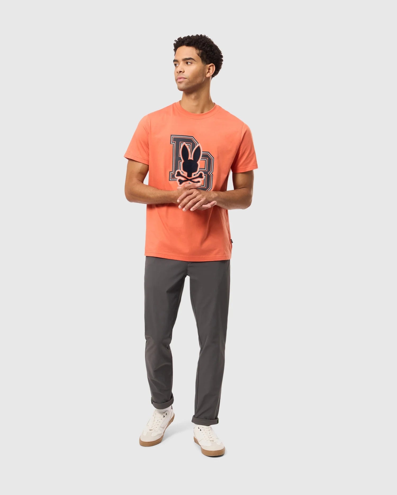 A young man stands against a plain background, wearing the Men's Zack Graphic Tee (B6U897D200) by Psycho Bunny, which features a Mickey Mouse design. He pairs it with gray pants and crisp white sneakers. Glancing to the side with his hands together, he effortlessly combines casual style with iconic charm.