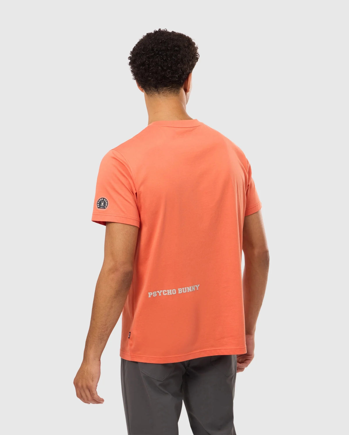 A man is sporting a coral MENS ZACK GRAPHIC TEE from Psycho Bunny, featuring the brand's name "PSYCHO BUNNY" prominently on the back. He faces away from the camera, highlighting his short, curly hair and complementing the look with gray pants.