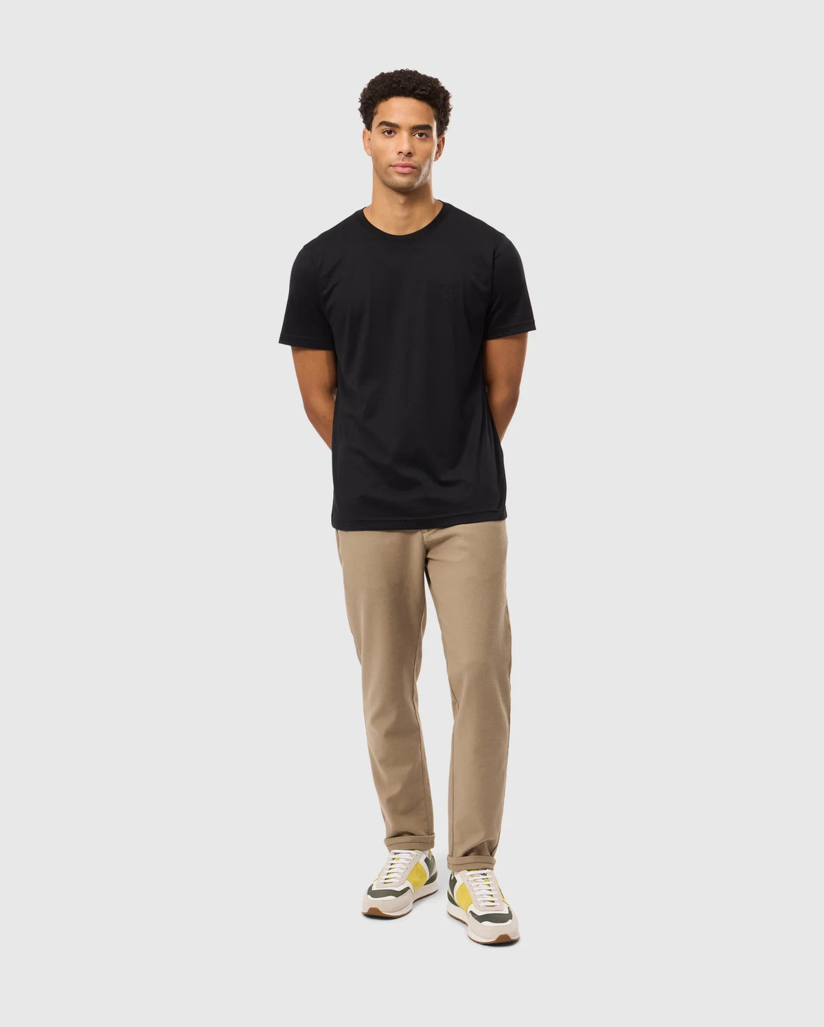 A person wearing the Psycho Bunny MENS JAMES PREMIUM CREW NECK TEE in black, made from high-quality Pima cotton, beige pants, and beige-yellow sneakers stands against a plain white background. Their hands are slightly tucked behind their back.