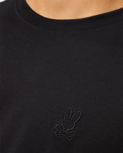 Close-up of a person wearing the Psycho Bunny MENS JAMES PREMIUM CREW NECK TEE - B6U952D200, a regular fit black T-shirt made from Premium Pima cotton, showcasing intricate embroidery of a rabbit's head above two crossed bones on the chest.
