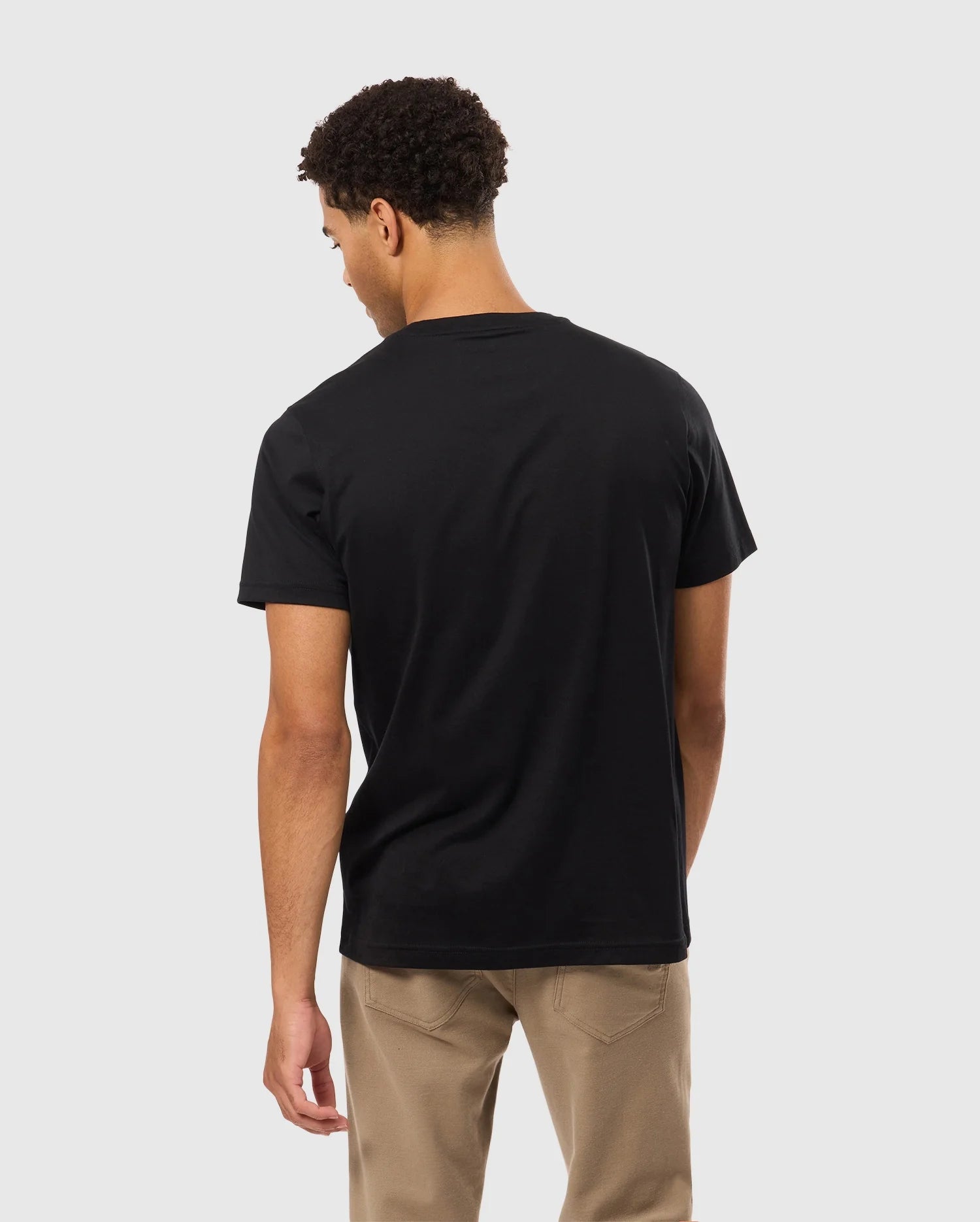 A person with curly hair is wearing a black Psycho Bunny MENS JAMES PREMIUM CREW NECK TEE - B6U952D200 made of Premium Pima cotton and beige pants, standing with their back to the camera against a plain gray background.