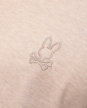 Close-up photo of beige fabric displaying an embroidered design on the Psycho Bunny MENS JAMES PREMIUM CREW NECK TEE - B6U952D200. The meticulous embroidery features a stylized bunny head above a pair of crossed bones, reminiscent of a skull and crossbones motif. The stitching is in a slightly darker shade than the premium Pima cotton fabric.