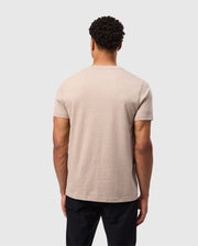 A person is standing facing away from the camera, wearing a beige Psycho Bunny MENS JAMES PREMIUM CREW NECK TEE - B6U952D200 made of premium Pima cotton and black pants. The background is plain and light gray.