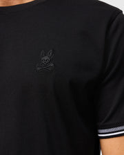 A person is wearing the Psycho Bunny MENS MALTON FASHION TEE - B6U966D200, a stylish black cotton tee that features a unique skull and crossbones emblem with rabbit ears on the chest. This men's fashion tee also showcases a striped edge on the sleeve, making it ideal for enhancing casual outfits.