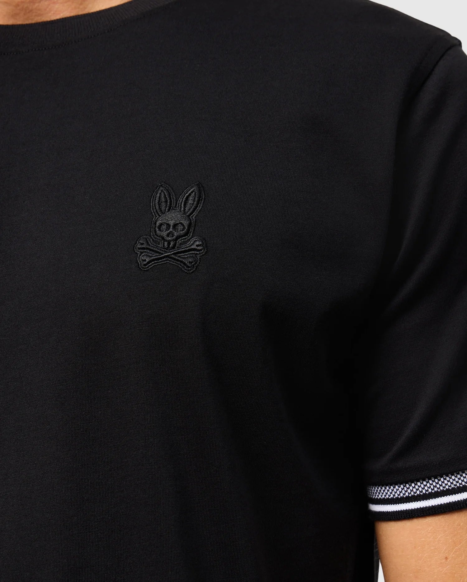 A person is wearing the Psycho Bunny MENS MALTON FASHION TEE - B6U966D200, a stylish black cotton tee that features a unique skull and crossbones emblem with rabbit ears on the chest. This men's fashion tee also showcases a striped edge on the sleeve, making it ideal for enhancing casual outfits.