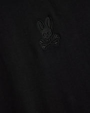 Close-up of the Psycho Bunny MENS MALTON FASHION TEE - B6U966D200, highlighting its stylish cotton fabric in black, adorned with an embroidered emblem resembling a skull and crossbones motif, featuring a rabbit head with long ears above crossed bones. The design is intricately stitched in subtle black thread, marking it as a standout piece in men's fashion.