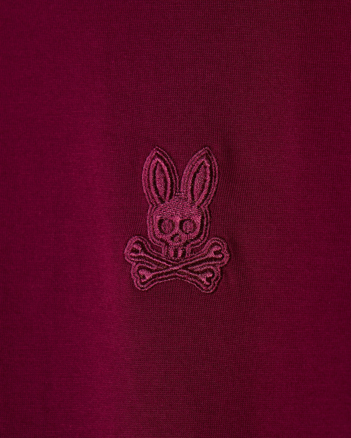 Detailed view of the maroon pima cotton fabric from the Psycho Bunny Men's Malton Fashion Tee (B6U966D200), featuring an embroidered logo of a bunny's head with crossed bones beneath. This stylish t-shirt showcases the logo stitched in a slightly darker shade of maroon for a subtle contrast, making it an excellent addition to any men's fashion tee collection.