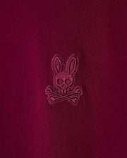 Detailed view of the maroon pima cotton fabric from the Psycho Bunny Men's Malton Fashion Tee (B6U966D200), featuring an embroidered logo of a bunny's head with crossed bones beneath. This stylish t-shirt showcases the logo stitched in a slightly darker shade of maroon for a subtle contrast, making it an excellent addition to any men's fashion tee collection.