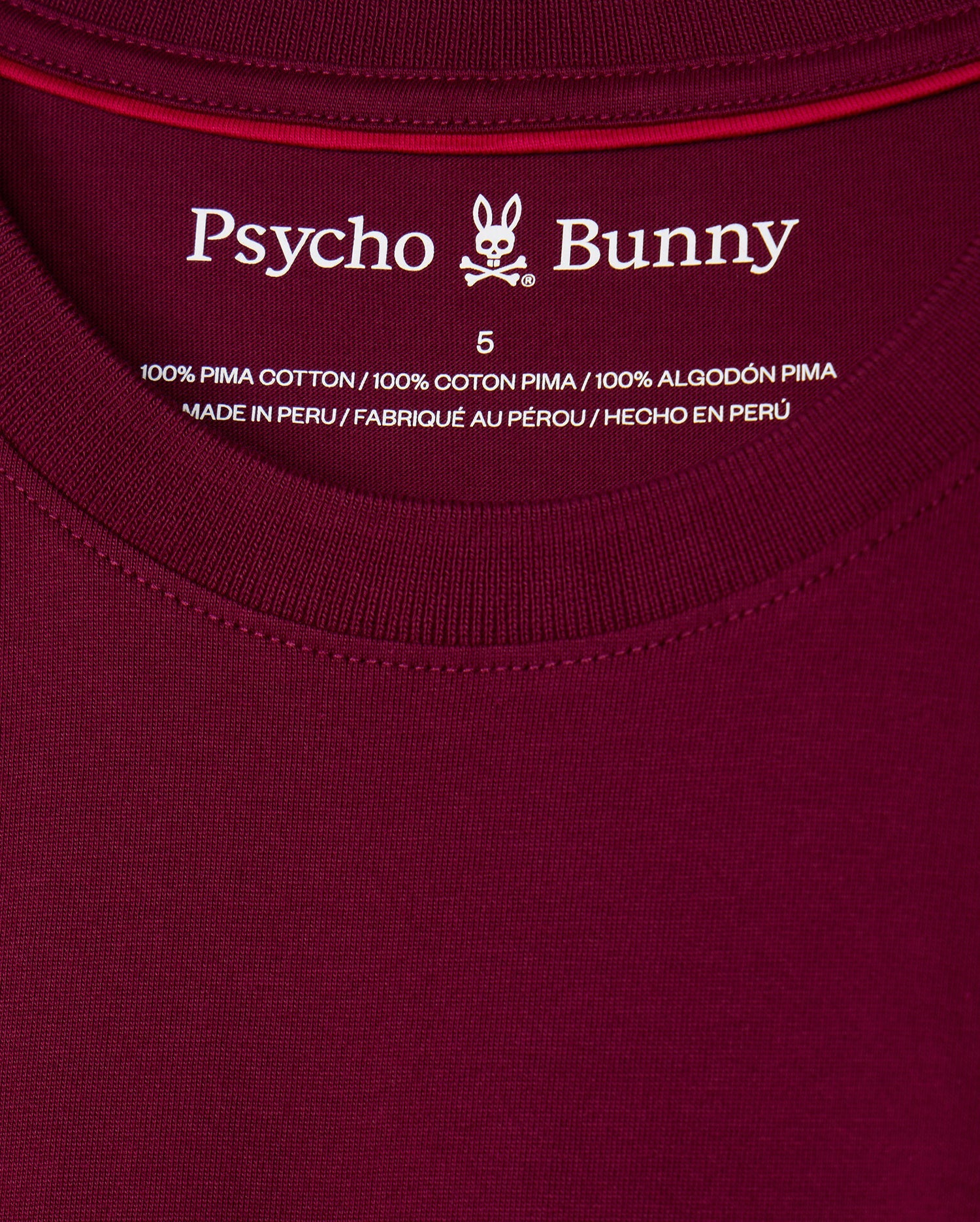 Close-up of the label on the maroon MENS MALTON FASHION TEE - B6U966D200 by Psycho Bunny, displaying size 5. The chic T-shirt label emphasizes "100% Pima Cotton" and notes that the shirt is "Made in Peru" in English, French, and Spanish.