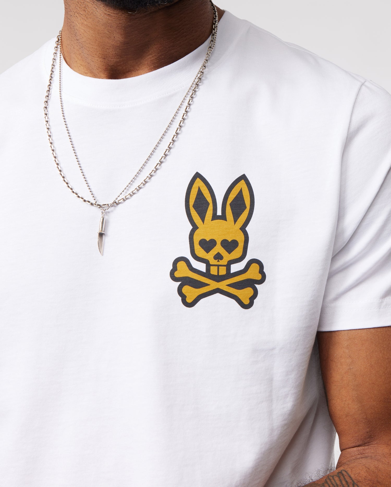 A person wearing a Psycho Bunny MENS VEGAS CITY TEE - B6U967X1PC made from soft Pima cotton jersey, featuring a yellow and black printed bunny logo on the chest. The person is also wearing a silver chain necklace with a pointed pendant. Only the torso and part of one arm are visible against a plain background.