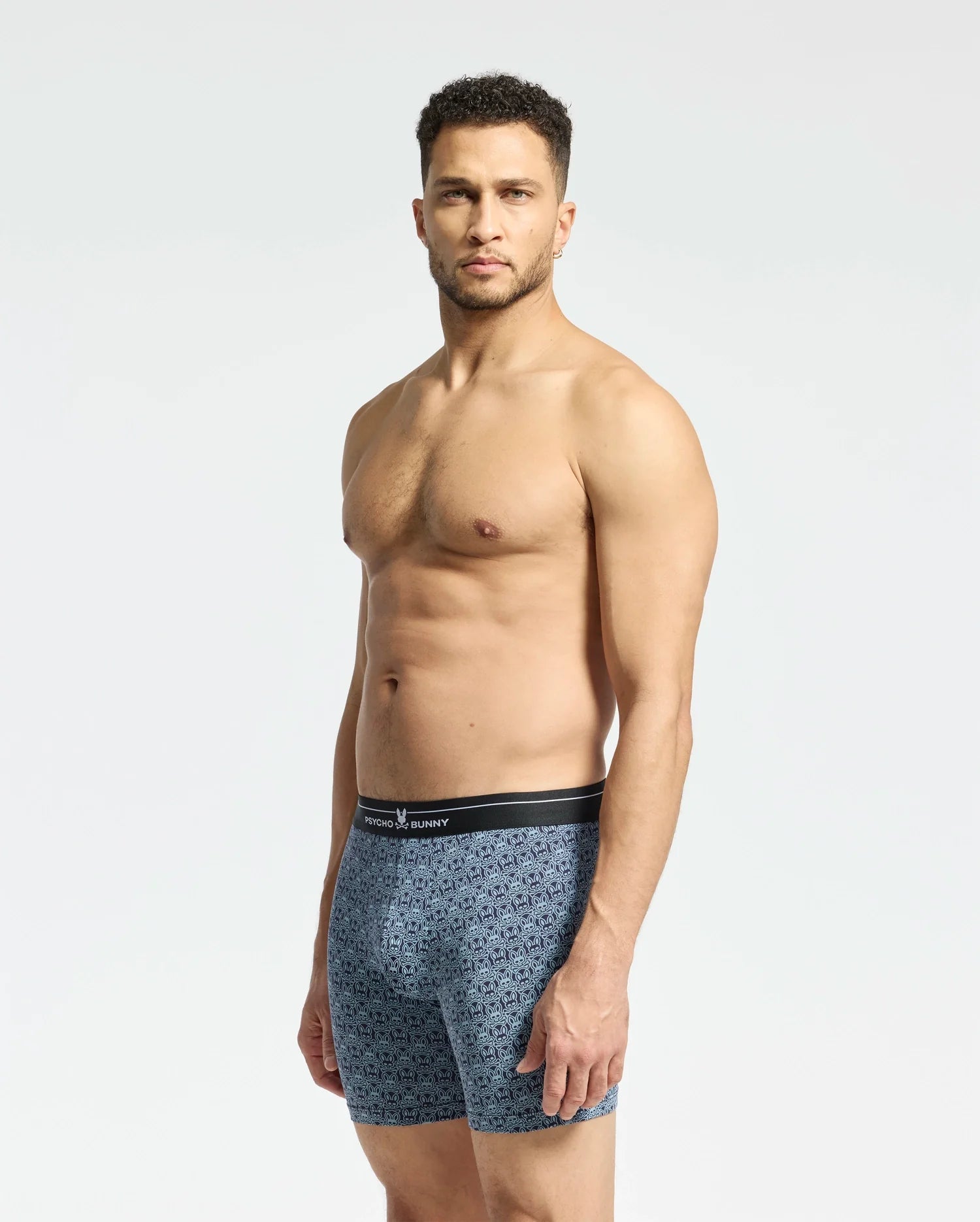 A man with short curly hair stands neutral, wearing blue patterned Psycho Bunny MENS 2 PACK BOXER BRIEF - B6V481F200. Crafted from soft-knit breathable stretch Pima cotton, the blue boxers feature a black waistband and offer comfort against a plain white background.