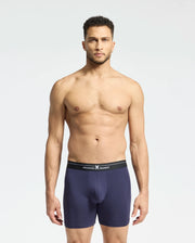 A man stands against a plain background wearing navy blue Psycho Bunny boxer briefs with a black waistband. Made from stretch Pima cotton, these breathable briefs promise supreme comfort. He looks directly at the camera with a neutral expression. Product: MENS 2 PACK BOXER BRIEF - B6V481F200.