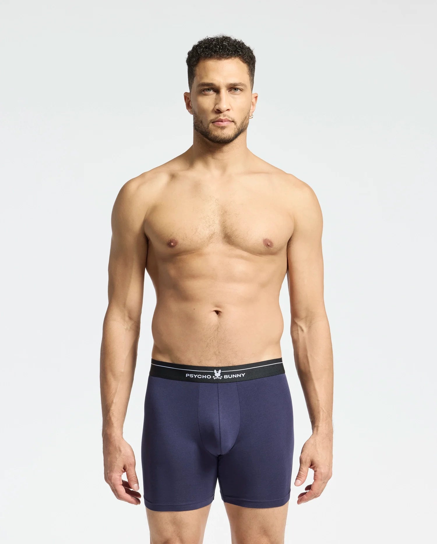 A man stands against a plain background wearing navy blue Psycho Bunny boxer briefs with a black waistband. Made from stretch Pima cotton, these breathable briefs promise supreme comfort. He looks directly at the camera with a neutral expression. Product: MENS 2 PACK BOXER BRIEF - B6V481F200.