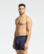 A man confidently stands in profile wearing Psycho Bunny's MENS 2 PACK BOXER BRIEF - B6V481F200 in dark blue Pima cotton with a black waistband. Set against a white background, his serious expression and relaxed posture highlight the breathable comfort and soft-knit fabric.