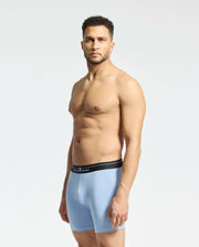 A man confidently poses against a plain light background, wearing Psycho Bunny's MENS 2 PACK BOXER BRIEF - B6V481F200. The light blue boxer briefs with a black waistband, crafted from stretch Pima cotton, perfectly complement his short curly hair and light beard as he gazes at the camera.