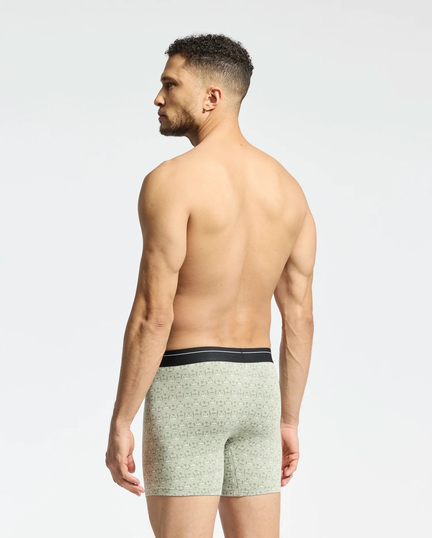 A man with short curly hair stands shirtless facing away, wearing Psycho Bunny's MENS 2 PACK BOXER BRIEF - B6V481F200 in light green with patterns and a black waistband. Made from a stretchy Pima cotton-Modal blend, they ensure all-day comfort against a plain white background.