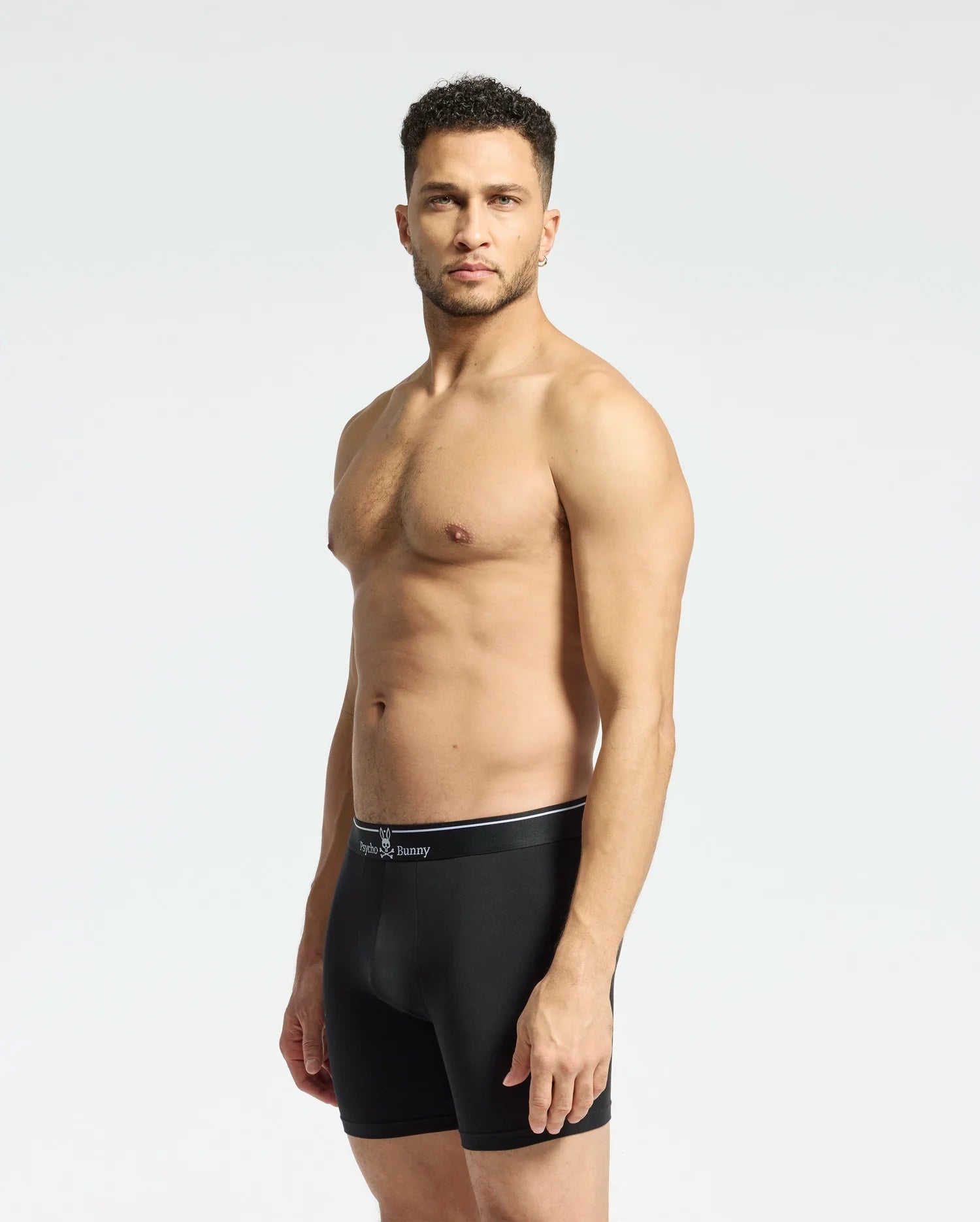 A man stands against a light gray background wearing the MENS SOLID KNIT 2 PACK BOXER BRIEF - B6V906ARCM by Psycho Bunny. He looks ahead with his arms at his sides, showcasing comfort and style in these ultrasoft modal-blend boxer briefs.