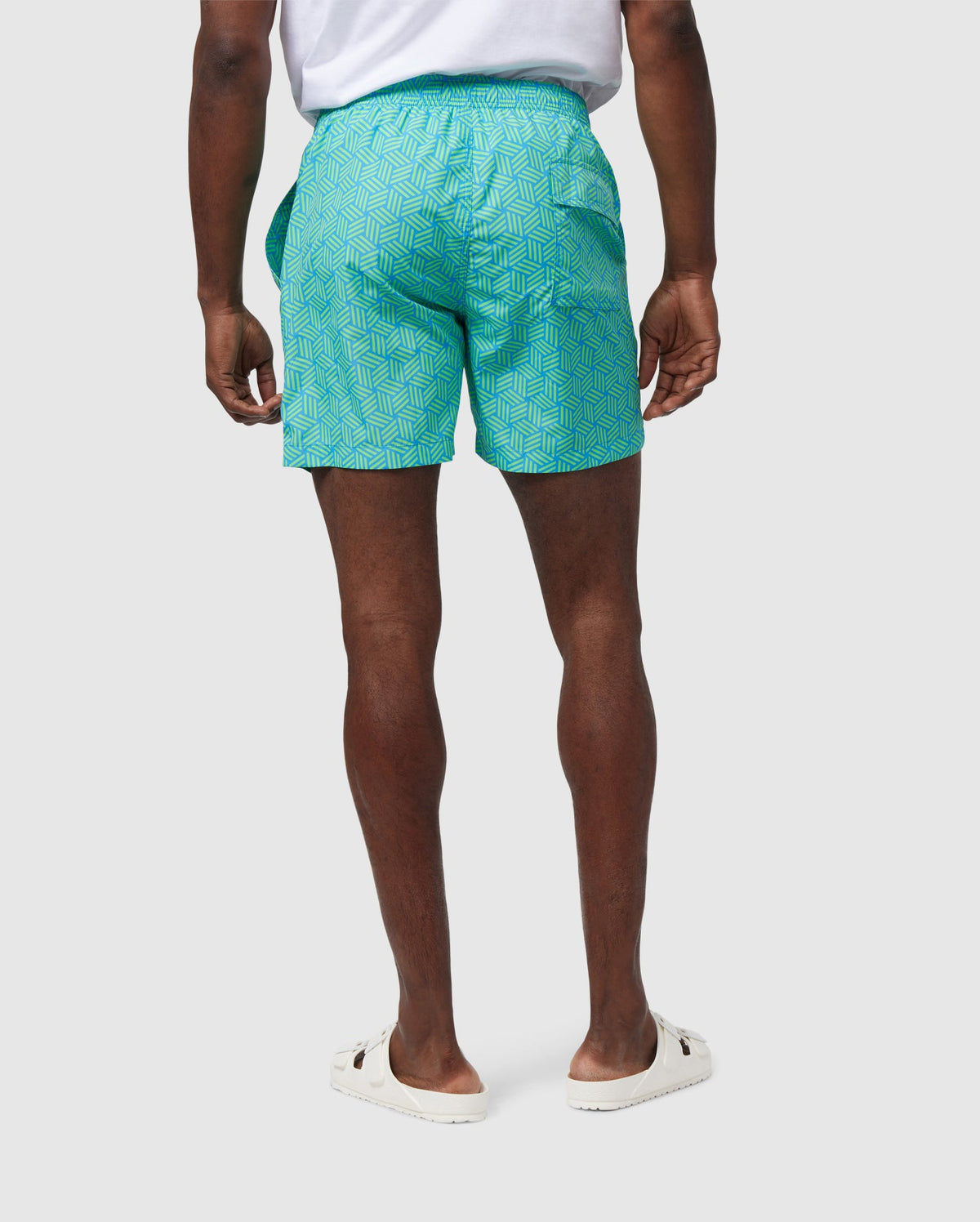 A man stands wearing a pair of teal geometric-printed pattern MENS JASPER LIGHTWEIGHT SWIM TRUNK - B6W168B2SW shorts and white slip-on shoes, pictured from the waist down against a white background.