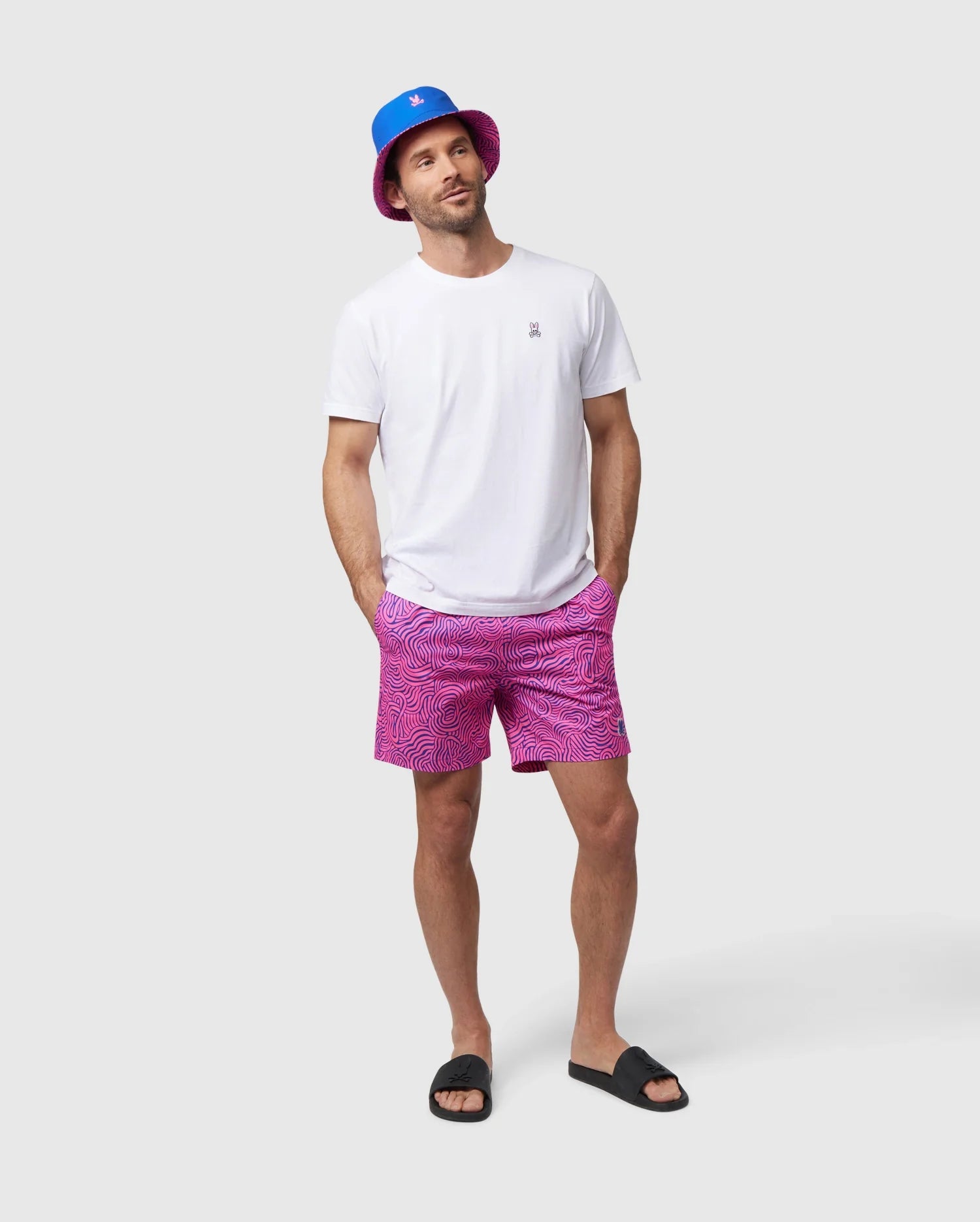 A person stands against a plain background, wearing a white t-shirt, Psycho Bunny VERONA PRINT SWIM TRUNK - B6W172B2SW with pink patterns, black slide sandals, and a blue bucket hat with a small pink logo. They have their hands in their pockets and a relaxed expression.