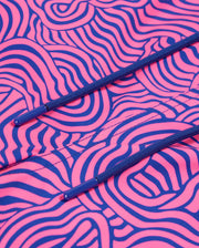 Close-up of a vibrant pink Psycho Bunny VERONA PRINT SWIM TRUNK - B6W172B2SW fabric with intricate black and blue swirly patterns. Two blue drawstrings with plastic tips are visible, positioned horizontally across the quick-dry fabric. The texture and design of the trunk's fabric are emphasized in the image.