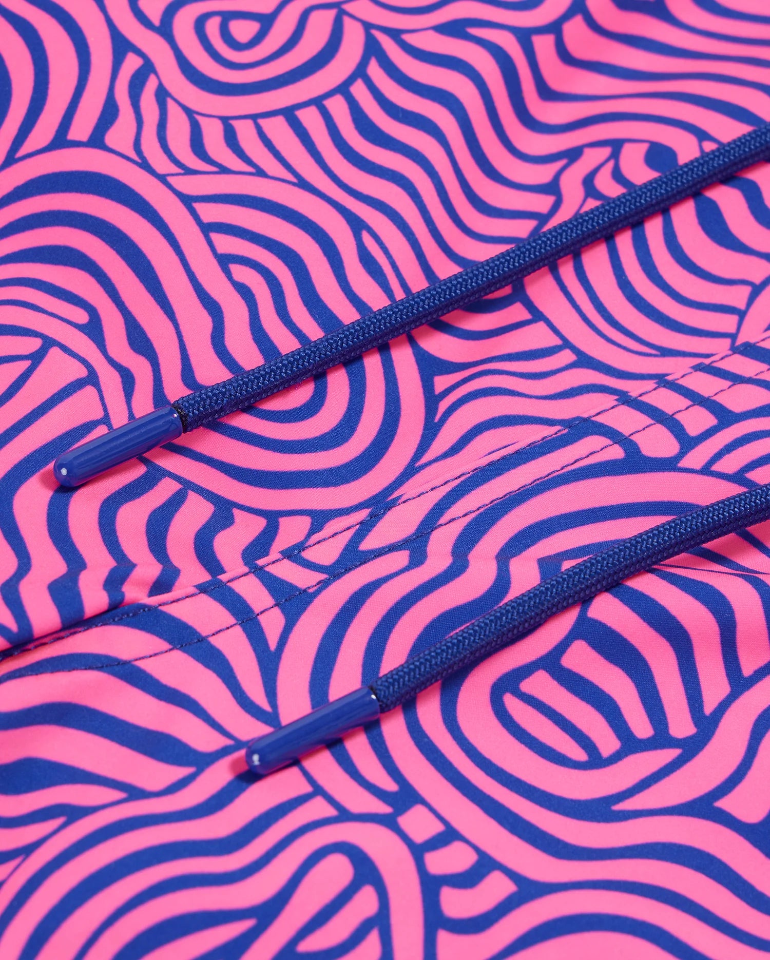 Close-up of a vibrant pink Psycho Bunny VERONA PRINT SWIM TRUNK - B6W172B2SW fabric with intricate black and blue swirly patterns. Two blue drawstrings with plastic tips are visible, positioned horizontally across the quick-dry fabric. The texture and design of the trunk's fabric are emphasized in the image.