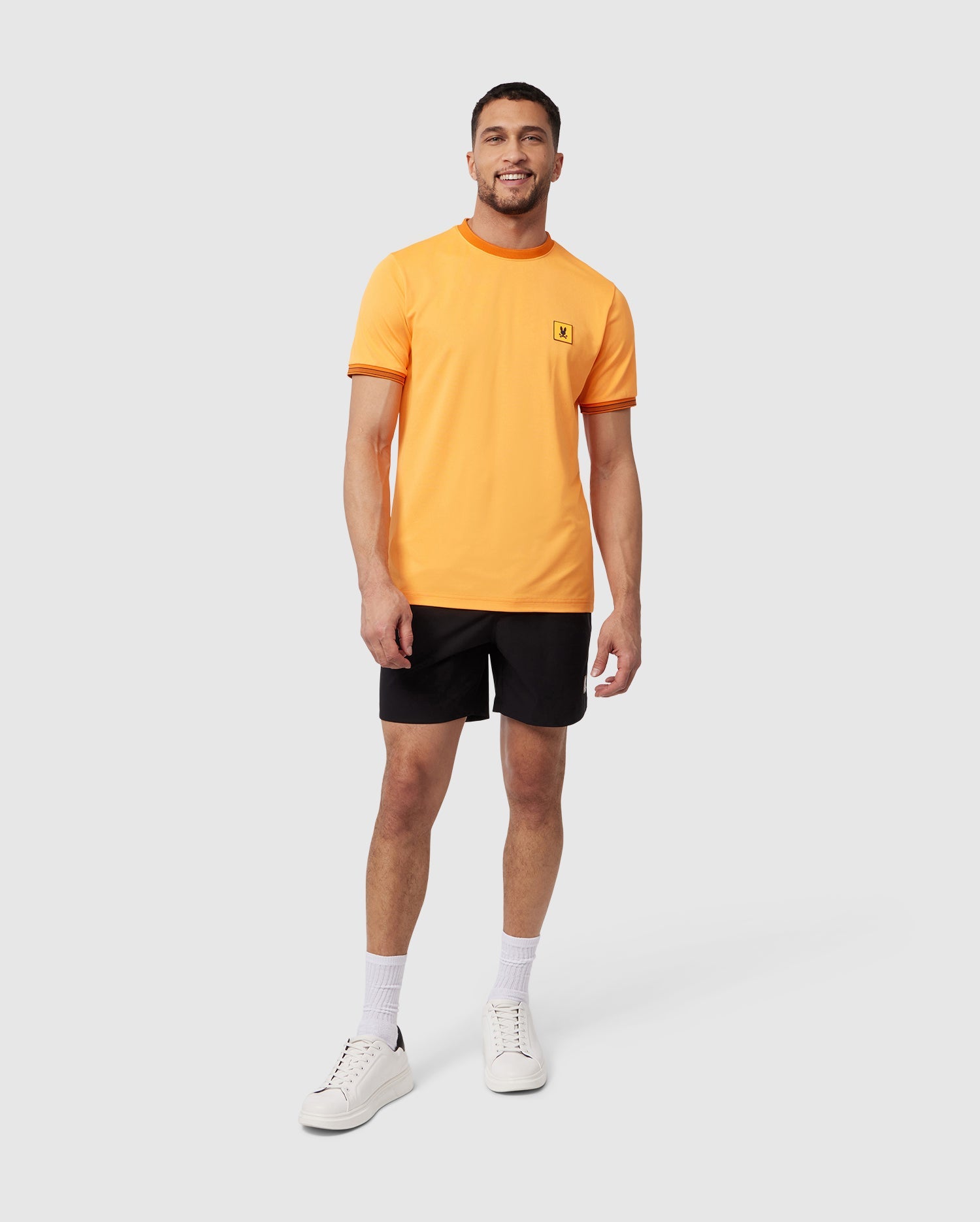 A smiling man standing, wearing an orange T-shirt, black Psycho Bunny MENS MALTA HYDROCHROMIC SWIM TRUNK, white socks, and sneakers against a plain white background.